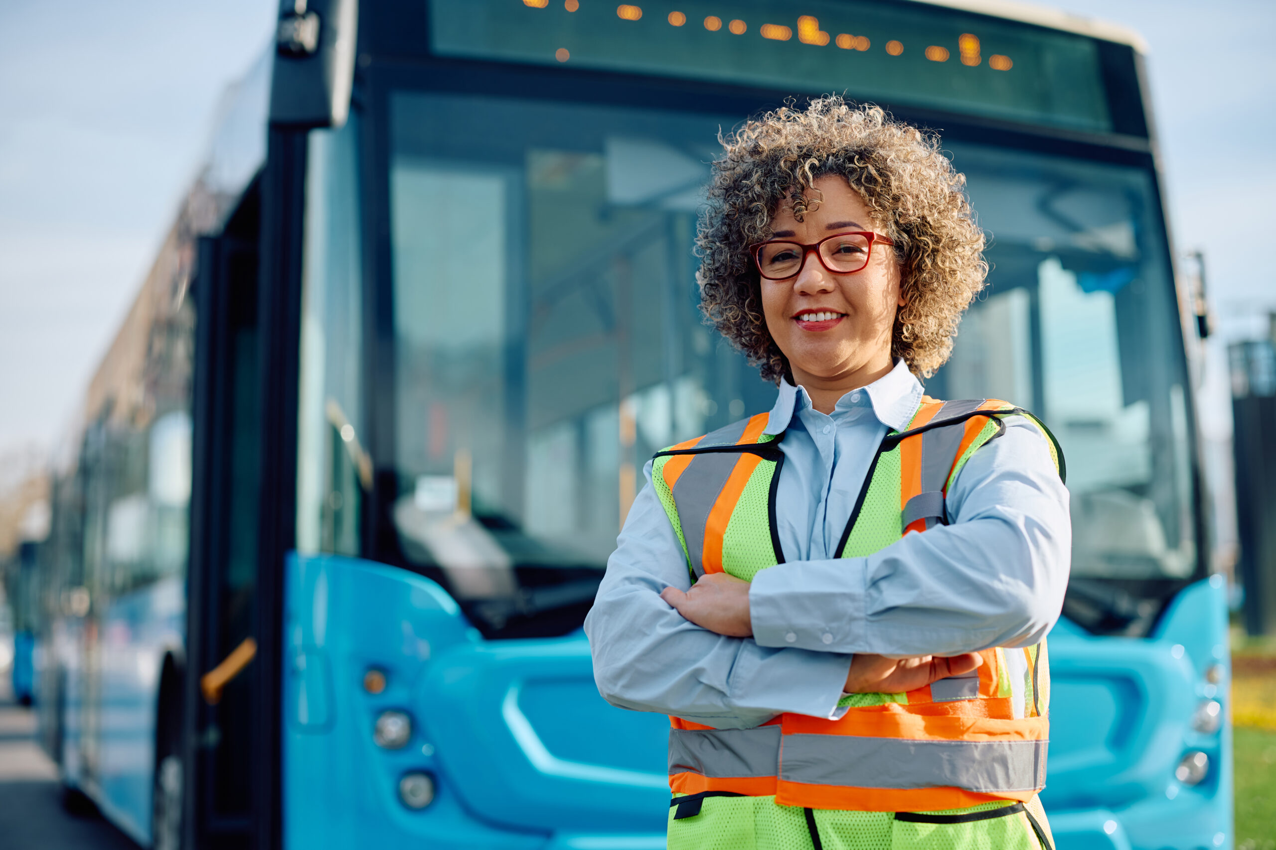 Happy bus driver standing with her arms crossed in front of a parked bus and looking at camera.